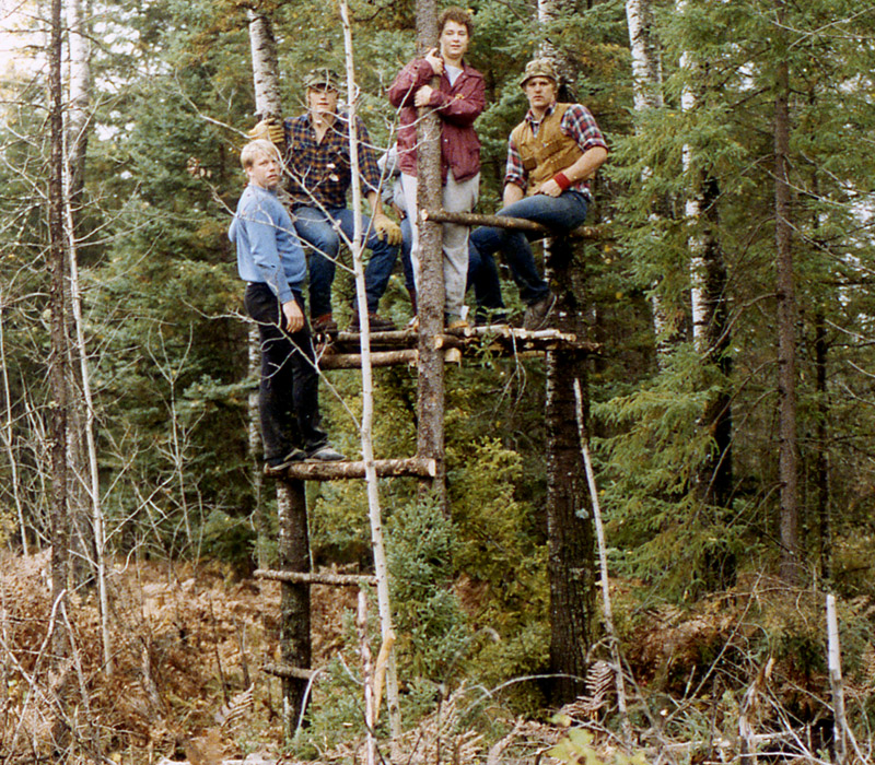Color photgraph of a much larger, and higher platform tree stand. There are 4 hunters from the Nordberg Hunting Group hanging on it.